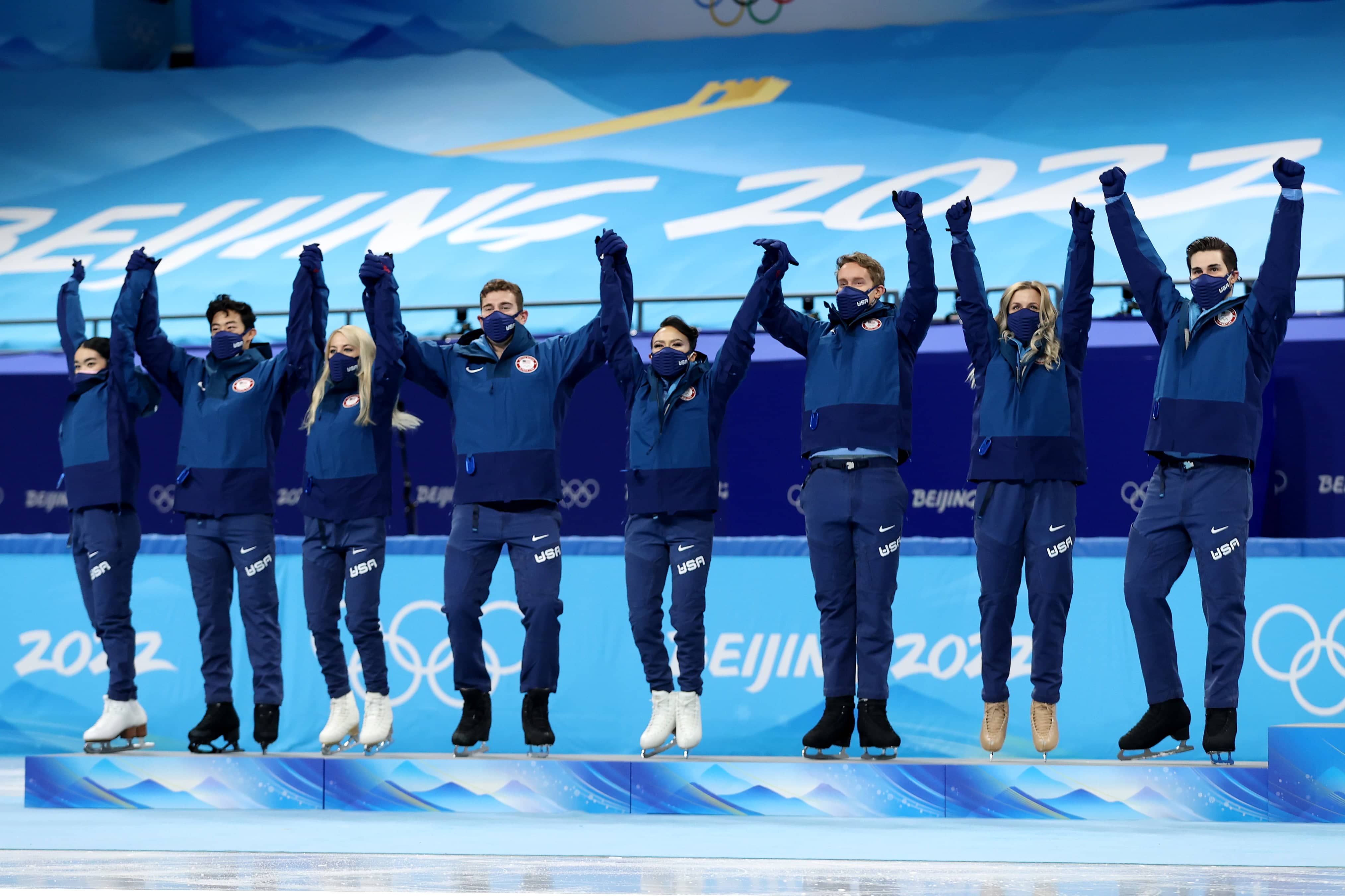 The nine members of the 2022 U.S. Team stand n the podium, holding hands with them up in the air in triumph. They wear blue jackets and pants.